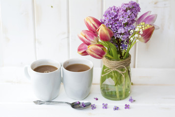 two cups of coffee with milk and bouquet of tulips and lilac on white wooden background