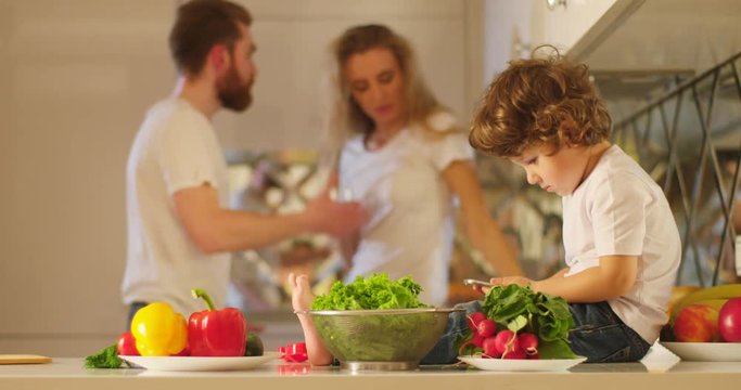 The little boy with the mobile phone in his arms is sitting on the kitchen while his parents are quarreling at the background.