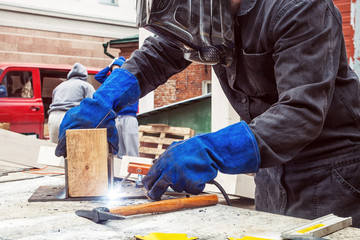 Close-up of a man in a black uniform, welding mask, protective gloves, welding a metal yellow...