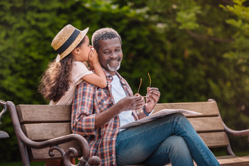 little adorable african american grandchild whispering to her smiling grandfather while sitting on bench in park - obrazy, fototapety, plakaty