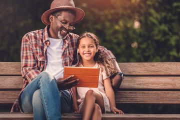 smiling african american grandchild and her grandfather listening music on digital tablet while...