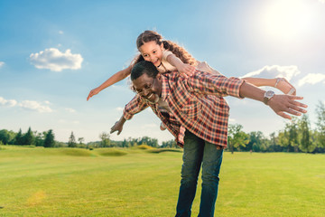 african american grandfather giving granddaughter piggyback ride on green lawn with sunlight