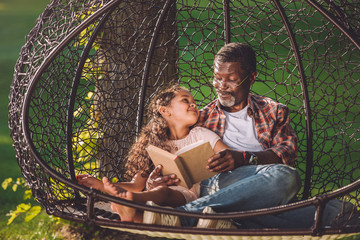 happy african american grandchild reading book while sitting in swinging hanging chair with...