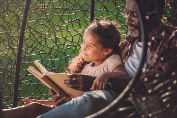 smiling african american girl reading book while sitting in swinging hanging chair with grandfather