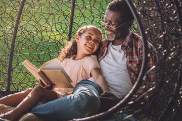smiling african american girl reading book while sitting in swinging hanging chair with granddad