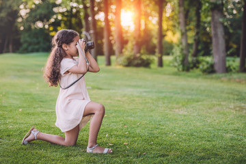 Fototapeta premium little african american girl taking photo on professional camera in park with backlit