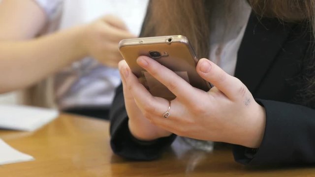 Schoolgirl dressed in a black suit sits at a school desk typing the text using a golden mobile phone. Close-up