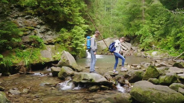 A young couple of tourists with backpacks crosses a mountain river in the forest