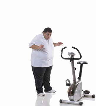 Full length of a shocked obese man looking at an exercise bike over white background 