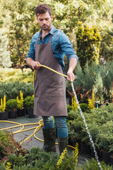 focused gardener in apron and rubber boots watering plants with sprinkler in garden