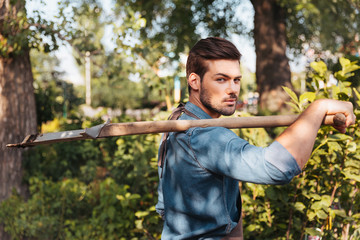 portrait of young gardener holding spade on shoulder and looking at camera in garden