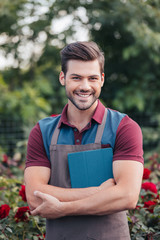 portrait of smiling gardener in apron holding digital tablet in hands while standing in garden