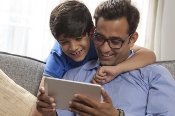 Smiling father using digital tablet sitting on sofa while his son looks from behind