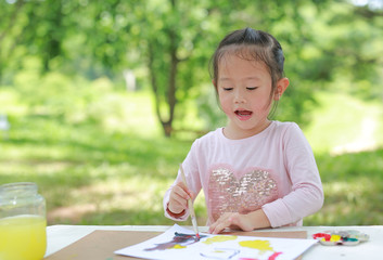 Asian child girl painting with paintbrush on arts paper on the table in the green garden.