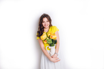 A studio shot of a girl standing and holding a white vase with flowers tightly. The girl has long wavy brunette hair and wears a yellow t-shirt and white skirt. Girl smiles widely.
