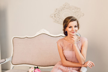 Charming blond girl sitting on a sofa. She has braided hair and holds her hand near chin. She wears peach color light dress and looks at the camera.
