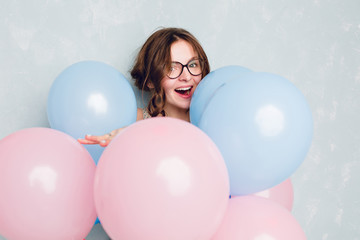 Close-up of cute brunette girl standing in a studio, smiling widely and hiding among blue and pink baloons. She wears black glasses, and has braided hair. She is having fun.