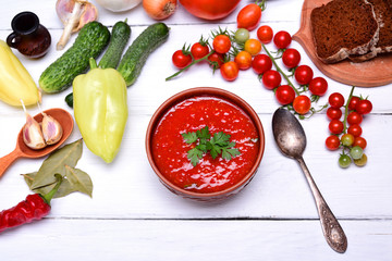 Soup gazpacho in a brown ceramic plate on a white wooden background, top view, around fresh vegetables and herbs for cooking