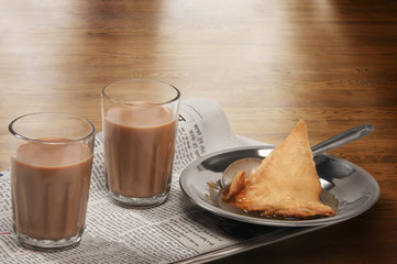 Glasses of chai on newspaper with plate of samosa over a wooden surface 