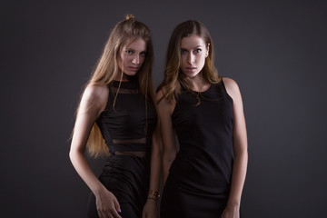 Two beautiful women in black night fashion dress posing on a gray background.