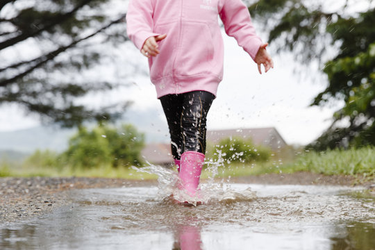 USA, Hide Park, Vermont. Little two year old blonde girl wearing a pink hooded sweater and pink rubber boots walks through puddles on a gravel road after heavy rain fall.