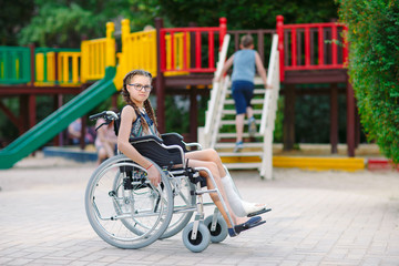 A girl with a broken leg sits in a wheelchair in front of the playground.