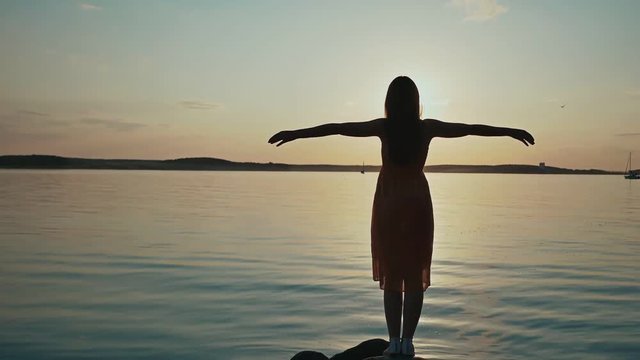 A young girl enjoys the sunset raising her hands and standing on the shore of the lake