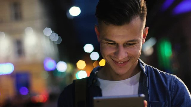 Young attractive man in the glasses typing in the tablet computer in the city at the night. Close up shot.