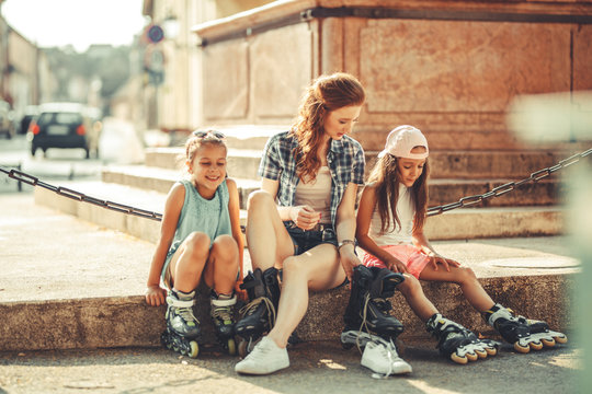 	Mother plays with her daughters on the street in neighborhood. They're sitting on stairs in city square and preparing to drive rollerblades. Family concept.	