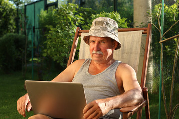 Portrait of old man in the garden sitting on a deck chair and holding a laptop in hands
