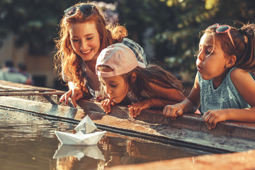 On a warm summer day, a mother, and her daughters joyfully set sail paper boats in a vibrant...