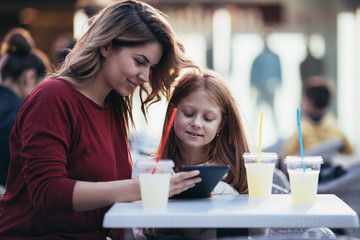 Mother and daughter sitting at cafe bar and looking something together at tablet.