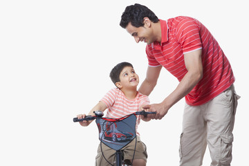 Father assisting son in riding bicycle over white background