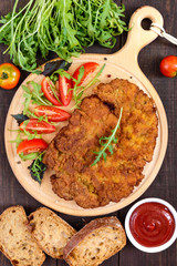 A large Viennese schnitzel  and tomato salad on a cutting board on a dark wooden background. Top view.