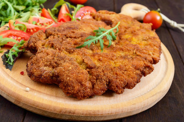 A large Viennese schnitzel  and tomato salad on a cutting board on a dark wooden background. Close...