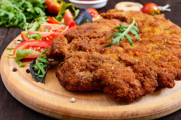 A large Viennese schnitzel  and tomato salad on a cutting board on a dark wooden background. Close...