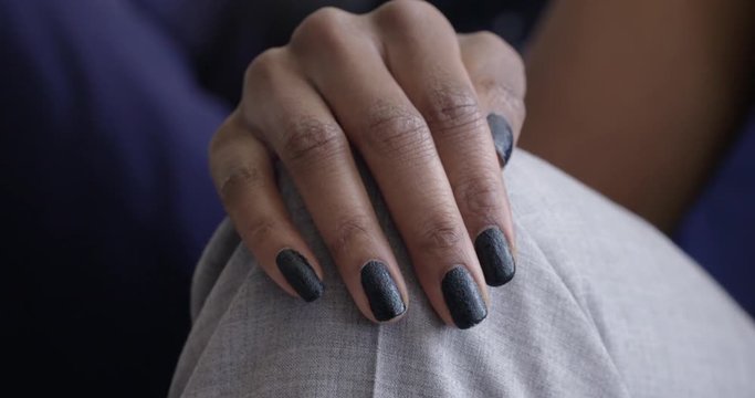 African American woman's hand with dark, glittering nail polish, moves up to rest on her knee in natural light. Slow motion 4K, recorded at 60fps.