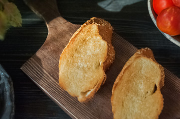 Two pieces of toasted bread. Toasted bread lies on a kitchen wooden board on a dark table. Bread with cheese, garlic, herbs and tomatoes.