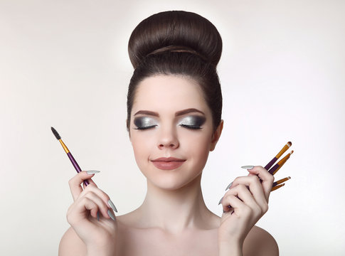Makeup artist. Pretty teen girl with cute bun hairstyle and fashion beauty makeup, brunette holding brushes in hand isolated on white studio background.