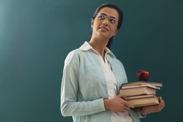 Thoughtful female teacher carrying stack of books and an apple against chalkboard 