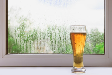 Glass of cold beer on the windowsill on a rainy summer day. The view from the window. Rain drops on glass. Holiday and vacation.