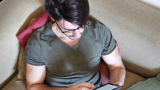 Handsome young man reading ebook on couch at home, wearing eyeglasses