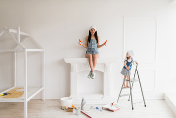 The young smiling mother does repair at home, she sits on fireplace and keeps thumbs up and her daughter stands on ladder and looks at mother