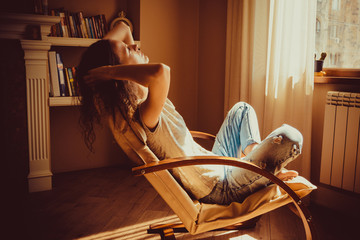 Woman relaxing after work in comfortable modern chair near window in livingroom. Warm natural light. Cozy home. Casual clothing. Casual style indoor. Room interior. Tired and thoughtful woman.