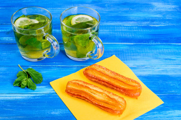 Two transparent cups with mint and lemon on a wooden blue background. And two cakes on a yellow paper napkin.