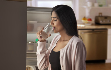 Young woman drinking water in front of open refrigerator 