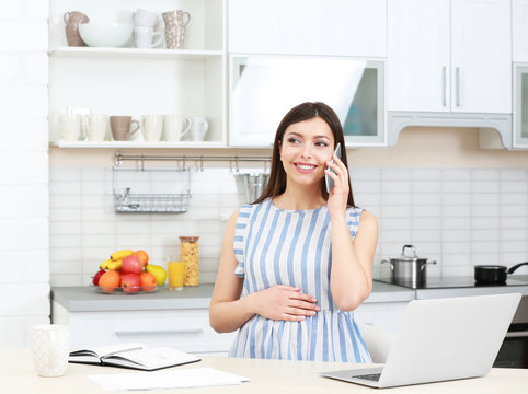 Young beautiful pregnant woman talking by phone while working in kitchen