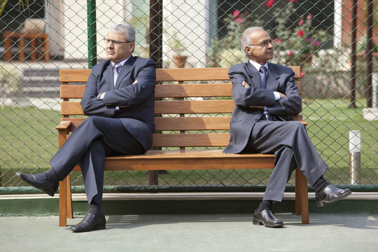 Senior male business executives looking away with arms crossed while sitting on bench in a tennis court 