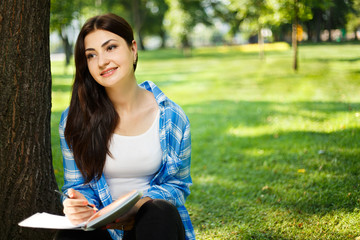 Female student taking notes in her notebook. Young smiling woman sitting in the park doing assignments. Campus life, education, inspiration concept
