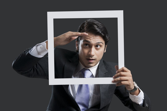 Young businessman shielding eyes while holding frame over black background